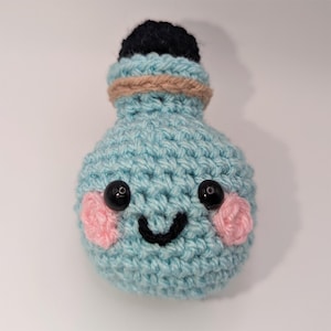 Puede incluir: Una botella de poción de ganchillo azul claro hecha a mano con una cara sonriente. La botella tiene una parte superior negra, mejillas rosadas y ojos de cuentas negras. Una banda de hilo marrón envuelve el cuello de la botella.