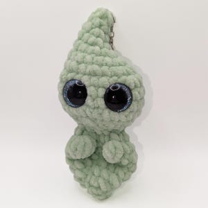 Puede incluir: Una criatura de ganchillo de color verde claro y afelpado con grandes ojos azules y negros brillantes. El artículo hecho a mano tiene una cabeza en forma de lágrima y brazos pequeños. Una cadena plateada está unida en la parte superior para colgar.