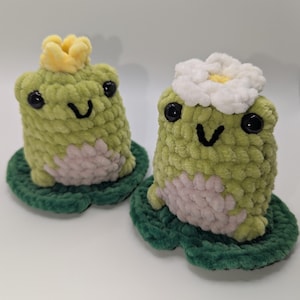 Puede incluir: Dos figuras de rana de ganchillo hechas a mano. Una rana lleva una corona amarilla, la otra una flor blanca. Ambas son de color verde claro con ojos negros y están sentadas sobre hojas de nenúfar verdes. Las ranas tienen vientre rosa claro.