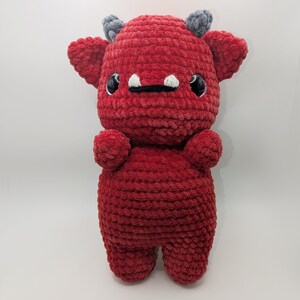 Puede incluir: Un monstruo de peluche rojo hecho a mano con cuernos grises, ojos negros y dientes blancos. El peluche tiene un aspecto texturizado y aterciopelado y está diseñado con rasgos redondeados. El peluche es una criatura linda y caprichosa.