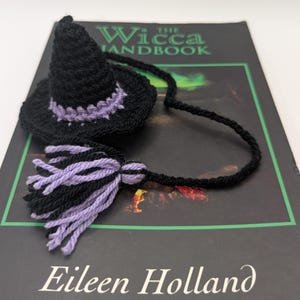 Puede incluir: Un marcapáginas de sombrero de bruja negro y morado tejido a ganchillo con una borla. El marcapáginas está sobre un libro titulado "The Wicca Handbook" de Eileen Holland. El sombrero tiene una parte superior puntiaguda y un ala ancha con un acento morado.