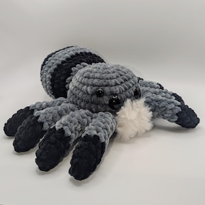 Puede incluir: Una araña de crochet suave, en tonos grises y negros. La araña tiene ojos grandes y negros brillantes y un mechón blanco y esponjoso. Las patas y el cuerpo están detallados con diferentes tonos de gris y negro. Juguete hecho a mano.