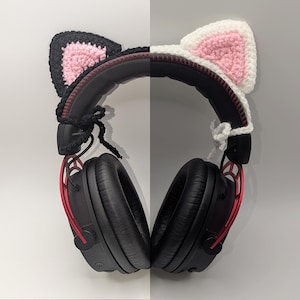 Puede incluir: Auriculares negros con detalles rojos, adornados con orejas de gato de ganchillo. Un juego de orejas es negro con rosa, el otro blanco con rosa. Los auriculares tienen una diadema y auriculares negros.