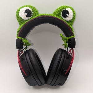 Puede incluir: Auriculares negros con una funda de oreja de rana de ganchillo verde. La funda de rana tiene ojos blancos y negros y un detalle de cremallera roja. Los auriculares tienen detalles en rojo.