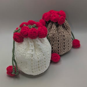 Puede incluir: Dos bolsas de crochet con cordón. Una es blanca, la otra es marrón. Ambas tienen pompones rojos alrededor de la parte superior y al final del cordón. La bolsa blanca tiene detalles verdes. Las bolsas son hechas a mano y tienen una apariencia texturizada.