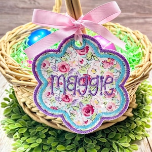 Puede incluir: Una etiqueta con estampado floral con el nombre "Maggie" bordado en morado, colgando de una cinta rosa atada a una cesta de mimbre. La etiqueta tiene un borde azul claro y morado. La cesta contiene huevos de Pascua.