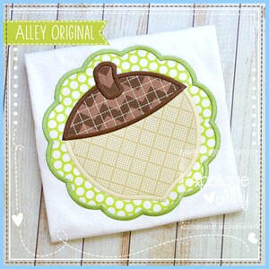 May include: A green and white polka dot applique with a brown and tan acorn design. The acorn has a brown cap with a chevron pattern and a tan body with a checkered pattern. The applique is surrounded by a scalloped edge.