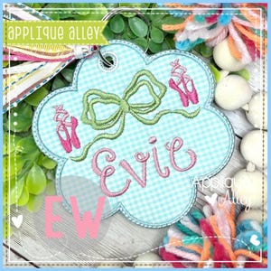 May include: A light blue, flower-shaped keychain with a gingham pattern, featuring a green bow, pink ballet shoes, and the name "Evie" in pink script. The keychain has a metal ring and a colorful tassel. The text "Applique Alley" is also visible.