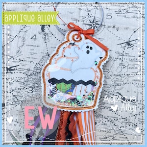 May include: A Halloween-themed shaker charm in the shape of a cupcake, featuring a white ghost and a green witch. The charm is decorated with orange, black, and white ribbons and the text "applique alley".