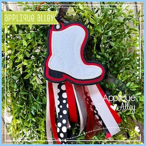 May include: A decorative applique featuring a cowboy boot design with a red, black, and white color scheme. The boot is light gray with a red outline and black heel. Ribbons in red, white, and black with polka dots hang below. The text "Applique Alley" is visible.