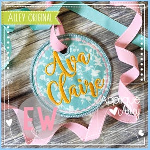 Può includere: Un'etichetta rotonda ricamata con il nome "Ava Claire" in oro. L'etichetta presenta un motivo floreale su sfondo azzurro, un nastro rosa e il testo "Alley Original".