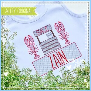 May include: A white baby onesie with a red embroidered design of a lobster trap with the word "SALE" on the sign and two lobsters. The onesie also has a red embroidered name tag with the name "ZAIN" on it.