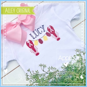 Puede incluir: Camiseta blanca con una guirnalda de langostas rojas y amarillas y el nombre "Lucy" bordado. La camiseta está sobre una superficie blanca con plantas verdes.