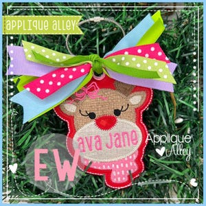 May include: A Christmas ornament featuring a reindeer design with the name "ava Jane" embroidered in pink. The ornament has a red border, a pink scarf, and a colorful bow with green, pink, and blue ribbons. The text "Applique Alley" is also visible.