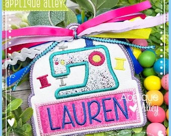 May include: A white, embroidered sewing machine-shaped ornament with the name "LAUREN" in pink and blue. The ornament has a teal outline and is adorned with colorful ribbons and the text "applique alley".