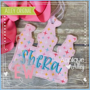 May include: A pink fabric crown-shaped ornament with the name "Shera" embroidered in blue. The crown has a colorful star pattern and is trimmed with pink stitching. The text "Alley Original" is visible.