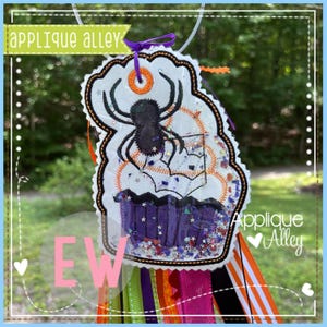 May include: A Halloween-themed tag with a spider and cupcake design. The tag is white with a black spider, orange accents, and purple cupcake. The tag has the text "Applique Alley" and "EW". Colorful ribbons hang from the bottom.
