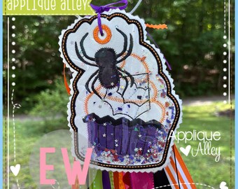 May include: A Halloween-themed tag featuring a spider and cupcake design. The white tag has a black spider, orange accents, and a purple cupcake. The tag has the text "Applique Alley" and "EW". Colourful ribbons hang from the bottom.