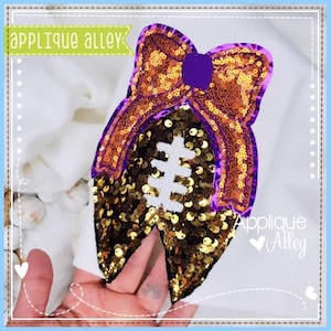 May include: A gold and purple sequined football with a purple bow on top. The football is on a white fabric background. The text "Applique Alley" is visible in the bottom right corner of the image.