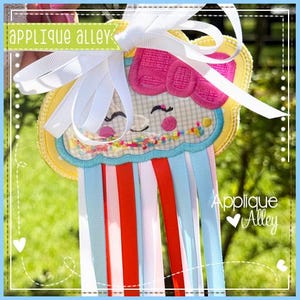 May include: A whimsical, handmade cupcake-shaped wall hanging with a smiling face, pink bow, and colorful sprinkles. It features a yellow and blue border, white ribbon, and red, white, and blue ribbons. The text "Applique Alley" is visible.
