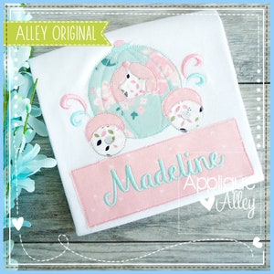 Könnte beinhalten: Ein weißes T-Shirt mit einem rosa und blauen gestickten Design eines Wagens mit einem Stern an der Seite. Der Name "Madeline" ist auf einem rosa Banner unter dem Wagen gestickt.