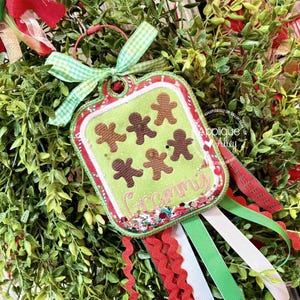 May include: A Christmas ornament with a green background featuring gingerbread men. The ornament has a red and green border, a green gingham bow, and the word "Grammy" embroidered on it. Red, white, and green ribbons hang from the bottom.