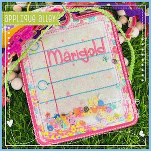 May include: A rectangular shaker tag featuring a clear vinyl center with the name Marigold embroidered in pink thread. The border is decorated with a colorful floral pattern and pink stitching. The bottom contains loose confetti pieces.