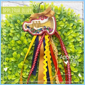 Pode incluir: Aplique decorativo com um design de trompete adornado com pedras preciosas coloridas e um laço dourado. Preso a fitas vermelhas, amarelas, azul marinho e douradas. O texto "Applique Alley" é visível.