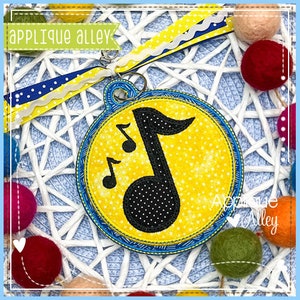May include: A yellow and blue musical note keychain with a black dotted musical note design. The keychain has a blue and yellow ribbon and the text "Applique Alley". The background is a white woven pattern with colorful felt balls.