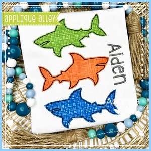 May include: White t-shirt featuring three colorful shark designs: green, orange, and blue. The name "Alden" is embroidered in black. The shirt is decorated with the text "Applique Alley".