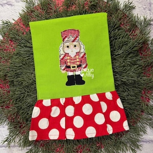 May include: A green Christmas-themed hand towel with a red and white polka dot ruffle. The towel features an embroidered nutcracker design in red, white, and black. The text "Applique Alley" is also embroidered on the towel.