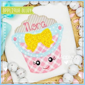 Pode incluir: Um item de tecido branco com um design de balde xadrez rosa, um laço de bolinhas amarelo e o nome "Nora" bordado em rosa. O balde tem um rosto sorridente e é adornado com detalhes azuis e marrons. O texto "Applique Alley" também é visível.