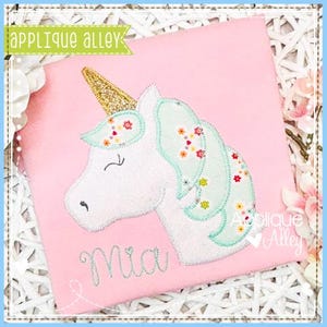 May include: A pink t-shirt with an embroidered unicorn applique. The unicorn has a gold horn, a white face, and a green mane with pink flowers. The name "Mia" is embroidered below the unicorn.
