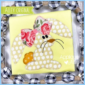 May include: Applique design of a bunny with a floral bow. The bunny is light brown with white polka dots and has a brown nose. The bow is pink and white with a floral pattern. The design is on a yellow background with the text "Applique Alley" in the bottom right corner.
