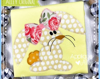 May include: Applique design of a bunny with a floral bow. The bunny is light brown with white polka dots and has a brown nose. The bow is pink and white with a floral pattern. The design is on a yellow background with the text "Applique Alley" in the bottom right corner.