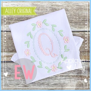 May include: A white shirt with a pink and green embroidered floral wreath and the letter Q in the center. The wreath is made of leaves and flowers. The letter Q is in a cursive font. The shirt is on a wooden background.