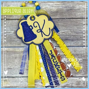 May include: A yellow and blue cheerleader mum with a cloud-shaped centerpiece featuring a blue megaphone and the letter "K". The mum has various ribbons, including one with the word "JACKETS" and football graphics. The text "Applique Alley" is visible.