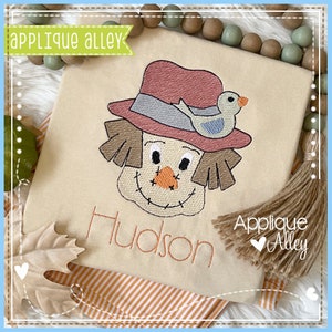 May include: Embroidered design of a scarecrow with a red hat and a bird perched on top. The scarecrow has a stitched smile and brown hair. The design is on a beige fabric with the name "Hudson" embroidered below.
