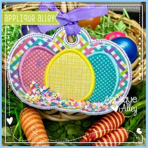 Puede incluir: Un aplique temático de Pascua con un diseño de huevo multicolor, con secciones rosas, amarillas y turquesas, acentuado con chispas y una cinta morada. El texto "Applique Alley" es visible.