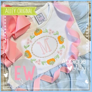 May include: White and light blue baby dress with a pink bow. The dress features an embroidered floral wreath with orange pumpkins and a pink initial "M". The text "Alley Original" and "Applique Alley" are visible.