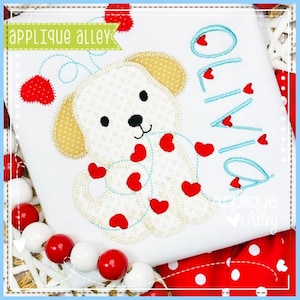May include: White t-shirt featuring an embroidered puppy with red heart accents and the name "Olivia" in turquoise. The puppy has tan ears and is surrounded by red hearts. The text "Applique Alley" is in a green banner.
