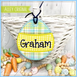 May include: A blue and yellow plaid Easter egg-shaped tag with a yellow gingham band that says "Graham". The tag is hanging from a white wicker basket.