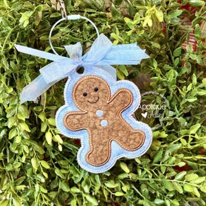 May include: A gingerbread man Christmas ornament with a light blue border and bow. The gingerbread man is brown with a smiling face and two small white buttons. The ornament hangs from a silver ring, set against a backdrop of green foliage.