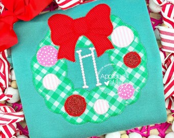 Op de afbeelding: Een turquoise shirt met een kerstkransontwerp. De krans is groen geruit met rode glitter- en roze stippenaccenten en een grote rode strik. De letter "H" staat in het midden. De tekst "Applique Alley" staat rechtsonder.