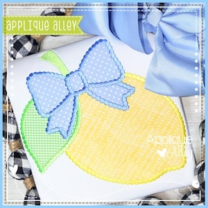 May include: A white fabric with a yellow lemon applique, a green leaf, and a blue bow with white polka dots. The lemon has a textured design and a scalloped edge. The text "Applique Alley" is visible.