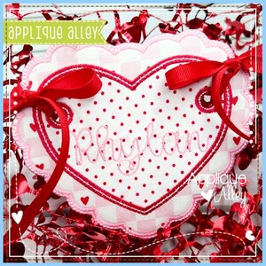 May include: A heart-shaped applique with the name "Rhylan" embroidered in pink script. The applique features a red polka dot center, a pink and white checkered border, and is adorned with red ribbons. The text "Applique Alley" is visible.
