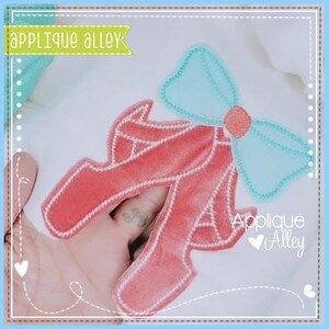 May include: A pink felt applique of a pair of ballet shoes with a light blue felt bow on top. The applique is on a white fabric background. The text "Applique Alley" is visible in the bottom right corner of the image.