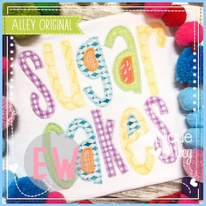 May include: A white t-shirt with the words "Sugar Cakes" in colorful applique letters. The letters are made of different fabrics, including gingham, polka dots, and stripes.