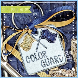 Puede incluir: Una etiqueta blanca bordada con las palabras "COLOR GUARD" en gris, con banderas cruzadas, una dorada y otra azul. La etiqueta está adornada con cintas azules y doradas y un anillo plateado. El texto "applique alley" está en la esquina superior izquierda.