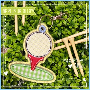 以下が含まれることがあります： 赤いティーと緑と白のチェック柄のベースが付いた刺繍されたゴルフボールのアップリケ。アップリケは、3本の木製ゴルフティーが付いたキーリングに取り付けられています。